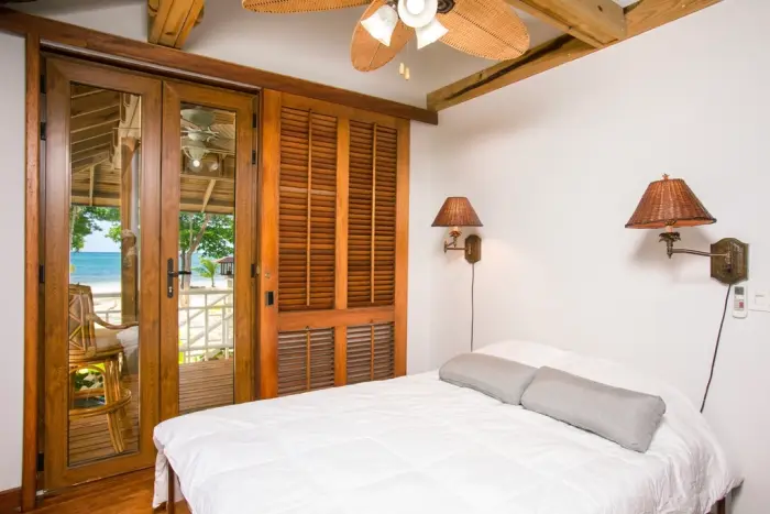 A white room with timber louvred doors, a ceiling fan and a glimpse of turquoise sea beyond the glass