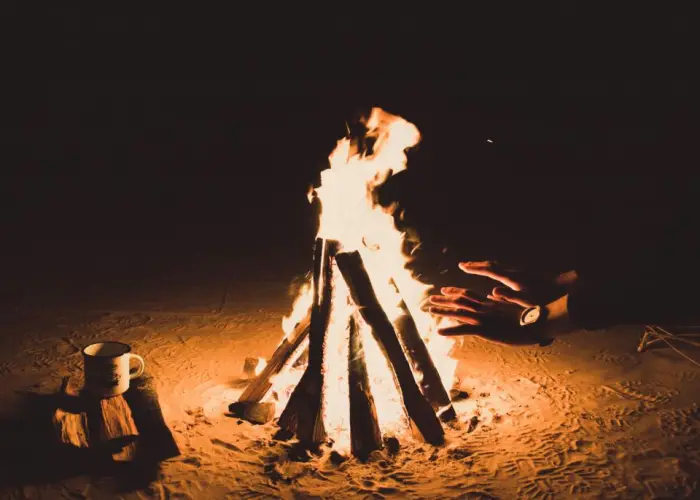 A campfire burning on sand at night beside a kettle and stacked logs