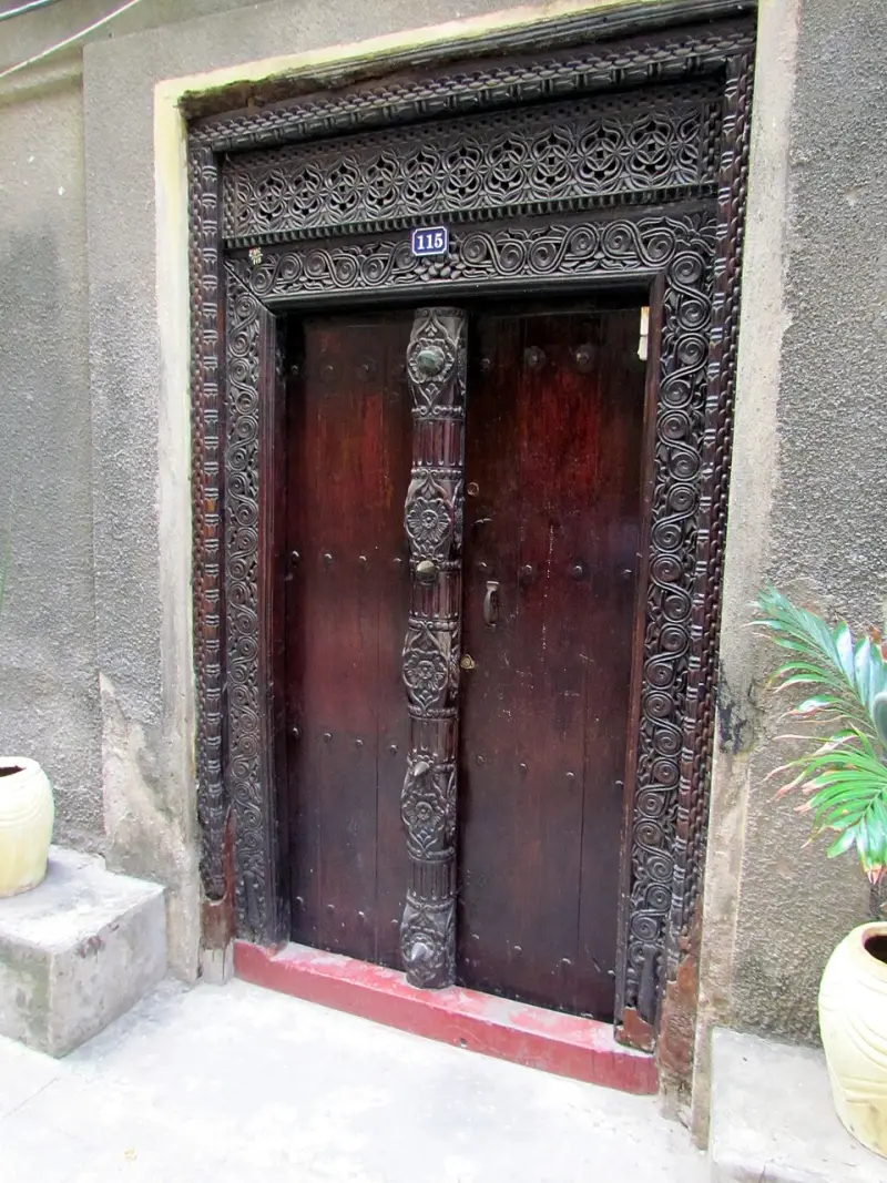 A tall carved wooden door with brass studs set in a white wall in Stone Town