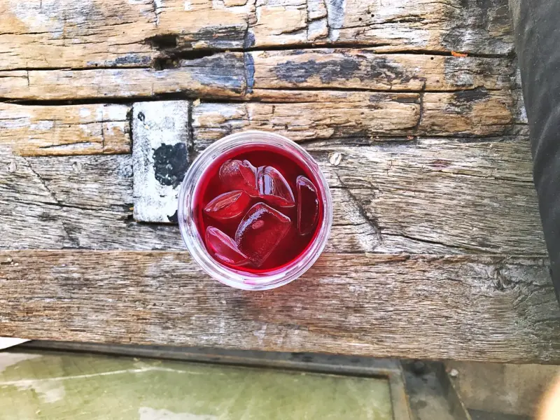 A glass of ruby hibiscus drink with ice on a weathered wooden table