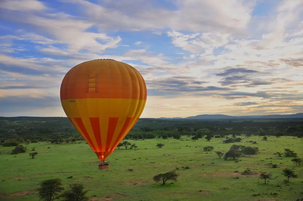An orange and yellow hot-air balloon drifting low over green savanna at sunrise