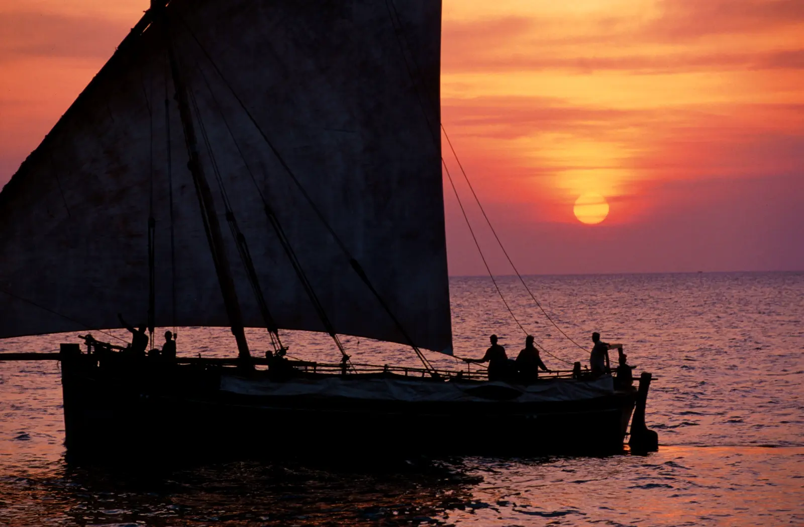 A dhow under full sail, silhouetted against an orange sunset over the sea