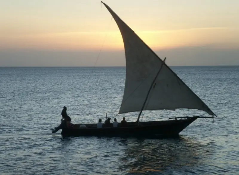 Guests aboard a dhow under a full white sail on a calm sea at dusk